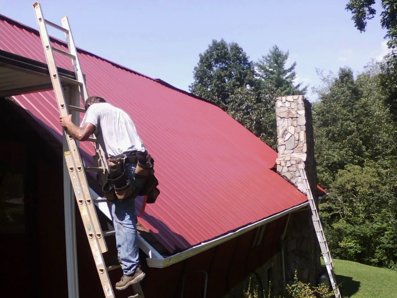 Roofer on a ladder beside a red metal roof and stone chimney — Soffit & Fascia Repair by Roofing Brothers in Redford