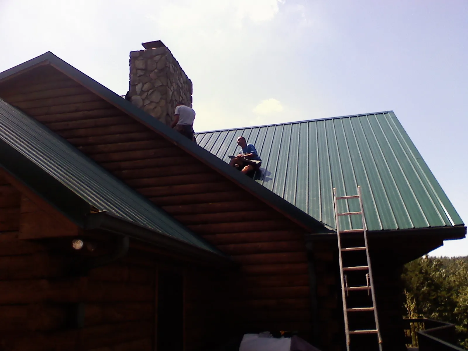 Roofers installing green metal panels beside a stone chimney on a log home in Redford